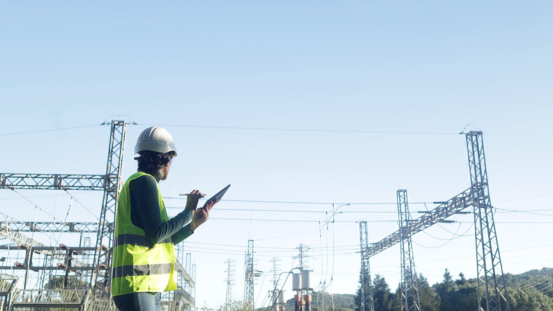 A field employee in high-visibility gear and hard hat reviews information on a tablet at a utility facility.